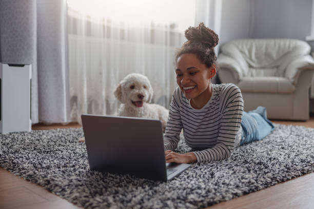 Happy positive female searching internet on laptop at cozy house on carpet, woman with dog at home